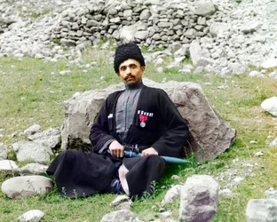 Dagesztáni típusok - Szunnita muszlim férfi, hagyományos ruhát és fejfedőt visel, oldalán tőrrel ül a földön, Orosz Birodalom, 1905 és 1915 között